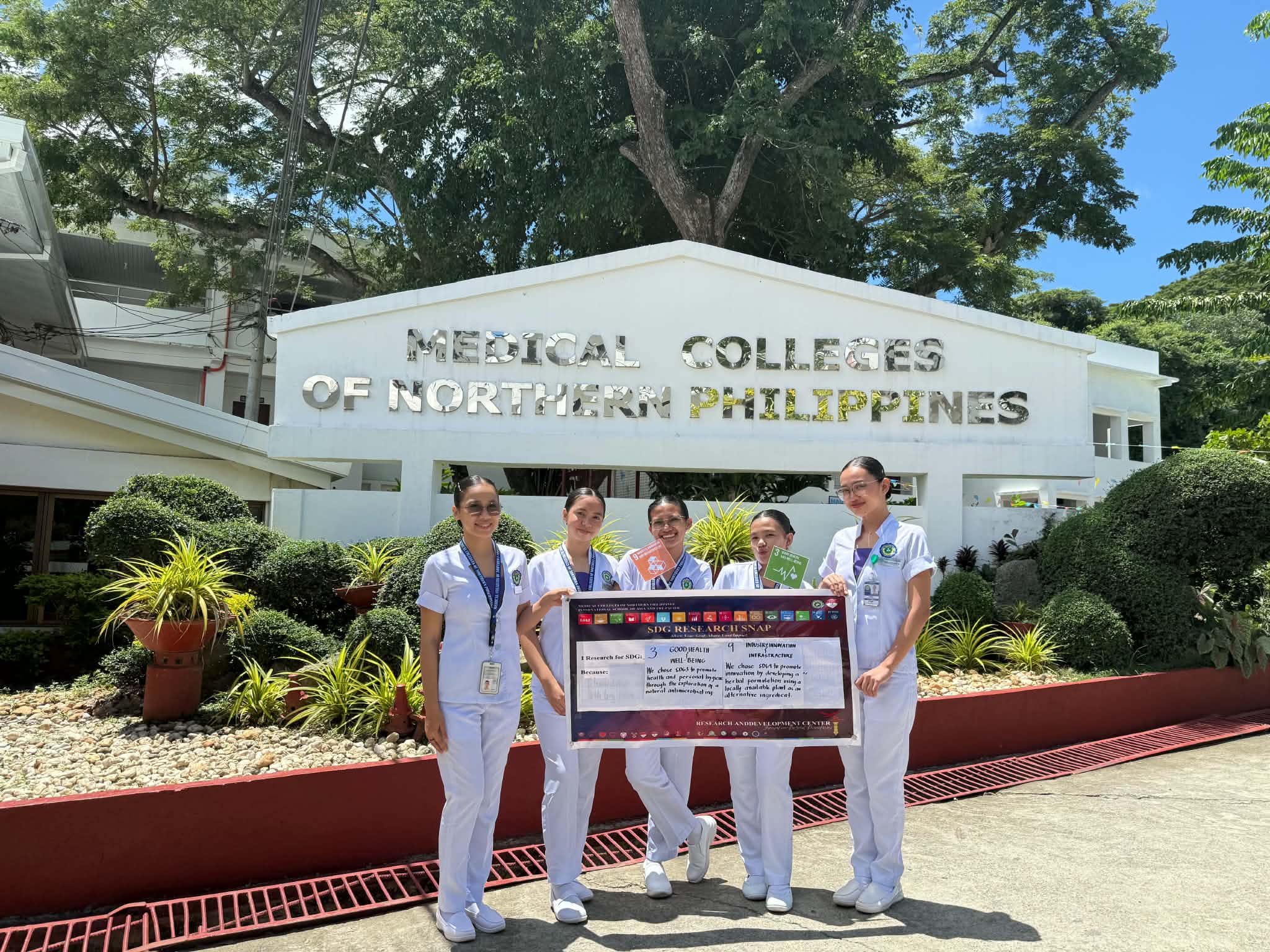 BS Pharmacy students holding SDG Research Snap banner at the Medical Colleges of Northern Philippines facade